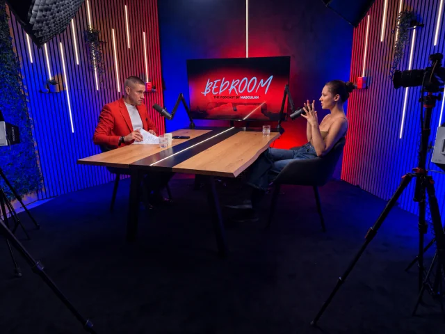 Podcast felvétel a StudioZone stúdióban: két szereplő a tölgyfa asztalnál, mikrofonokkal, piros-kék fényben, kamerákkal